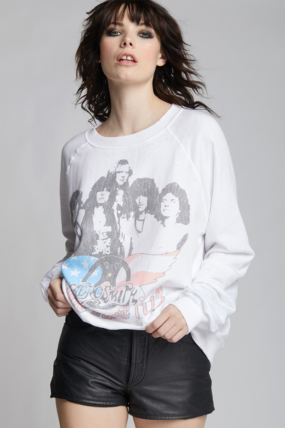 A woman modeling a white long-sleeve crewneck sweatshirt with a vintage Aerosmith Live band graphic on the front, paired with black leather shorts, against a solid gray studio background.