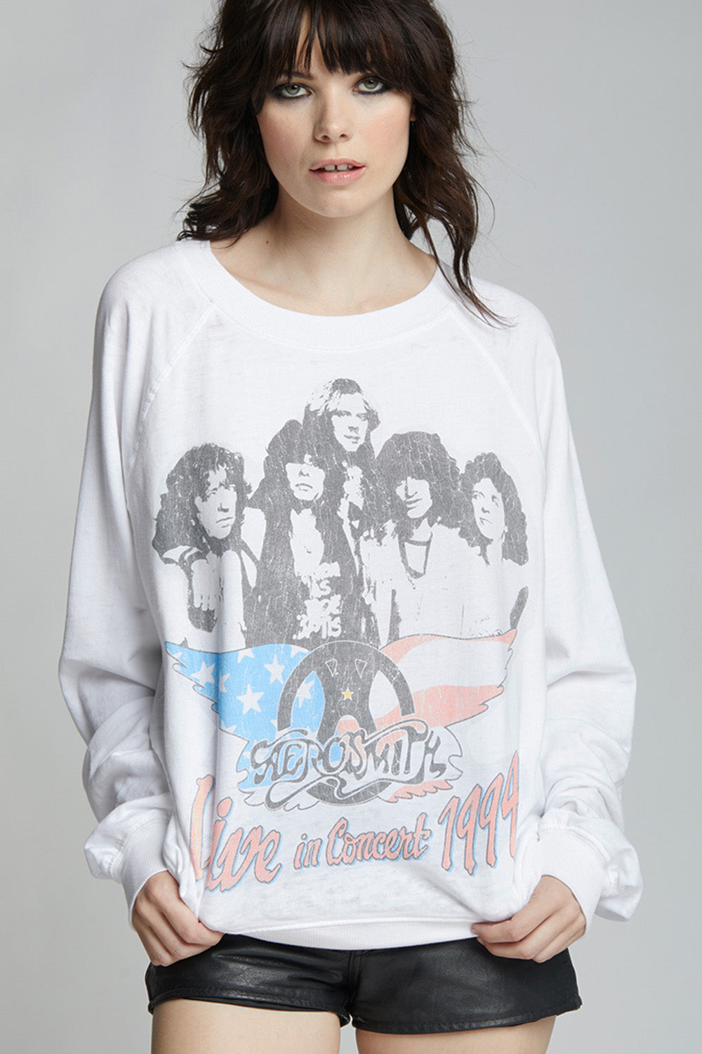A woman wearing a white long-sleeve graphic sweatshirt featuring the band Aerosmith and the text 'Live in Concert 1994' with a peace sign, paired with black shorts, against a light gray studio background.