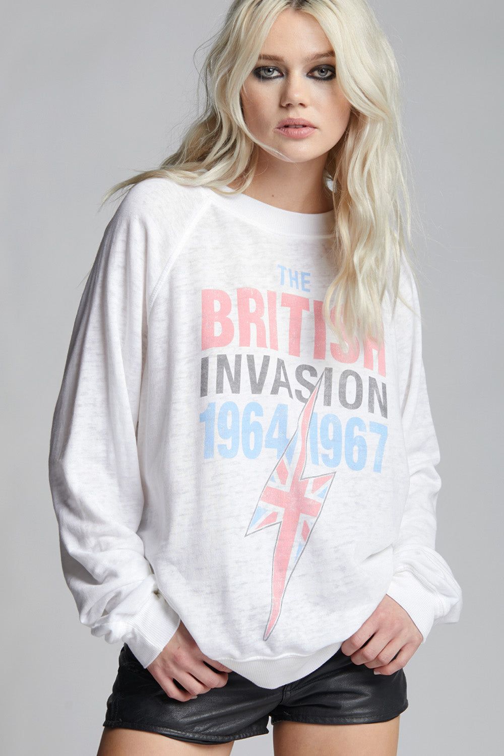 A blonde model wearing a white crewneck sweatshirt with a graphic print that reads 'THE BRITISH INVASION 1964 1967' in red, gray, and blue, alongside a Union Jack lightning bolt design, paired with black leather shorts against a gray studio background.