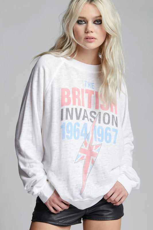 A blonde model wearing a white crewneck sweatshirt with a graphic print that reads 'THE BRITISH INVASION 1964 1967' in red, gray, and blue, alongside a Union Jack lightning bolt design, paired with black leather shorts against a gray studio background.