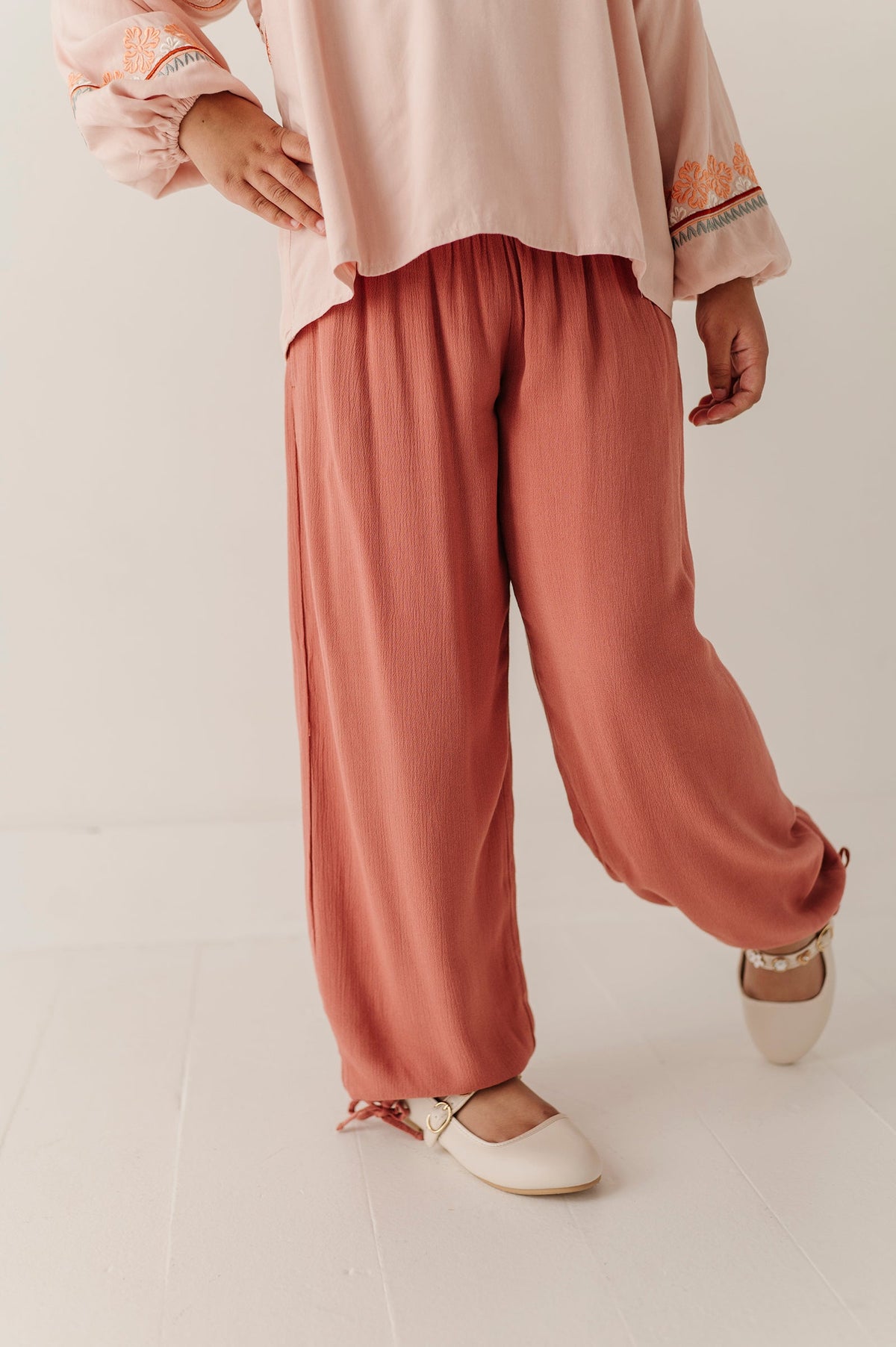 Person wearing a light pink top and Brick Dust-colored harem tie pants on a white background