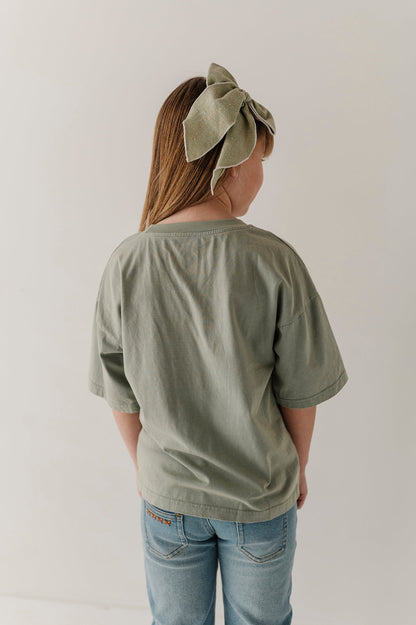 A child viewed from behind wearing an oversized sage green t-shirt and light blue jeans, with a matching sage green fabric bow headband. The photo is shot against a plain, light off-white background.