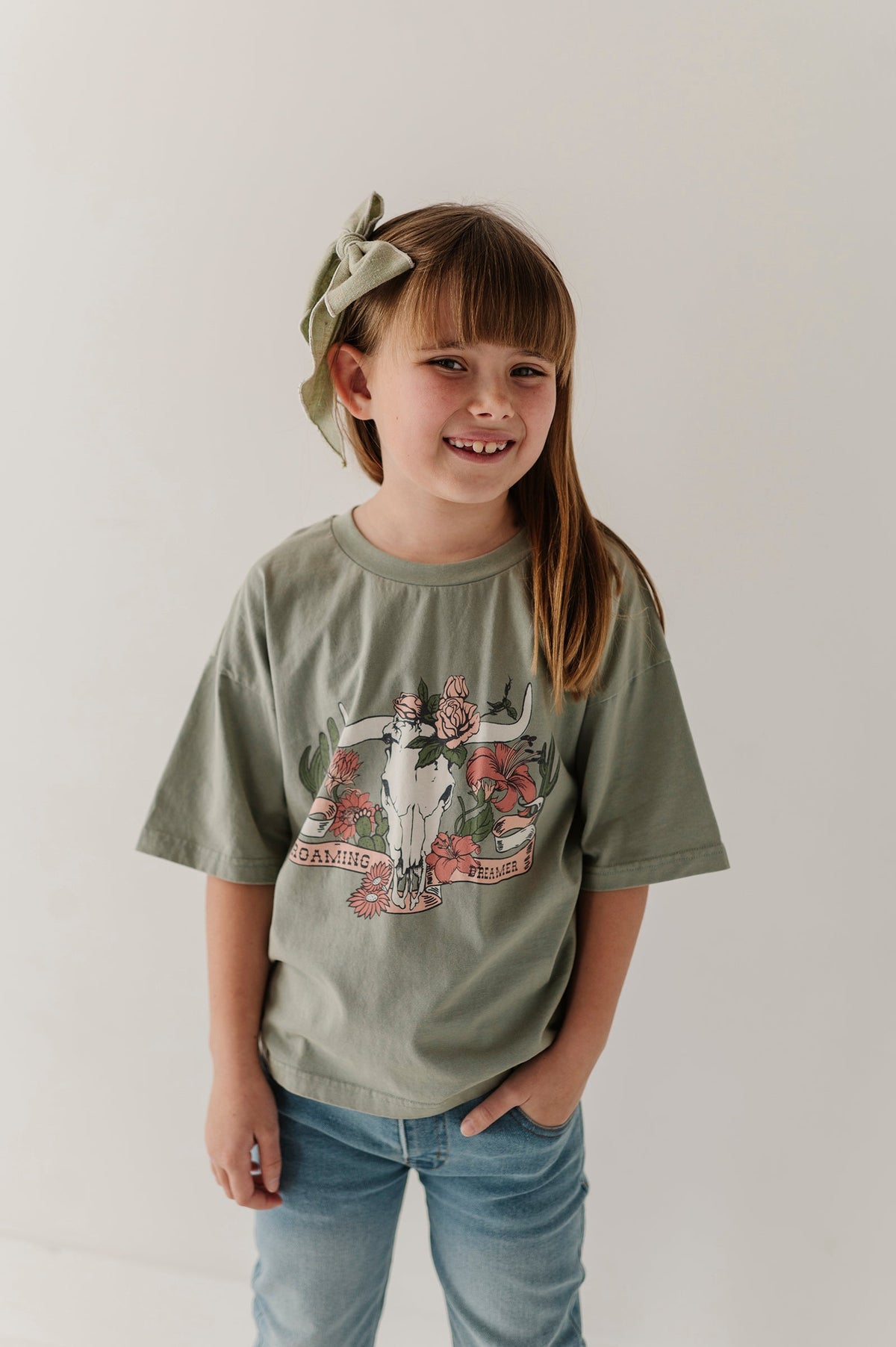 A young girl with a green bow headband smiles while wearing an oversized sage green graphic "Roaming Dreamer' t-shirt and blue jeans against a plain white background.