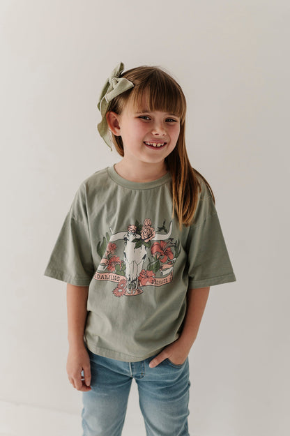 A young girl with a green bow headband smiles while wearing an oversized sage green graphic "Roaming Dreamer' t-shirt and blue jeans against a plain white background.