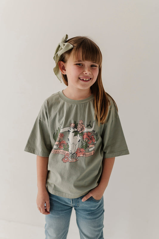 A young girl with a green bow headband smiles while wearing an oversized sage green graphic "Roaming Dreamer' t-shirt and blue jeans against a plain white background.