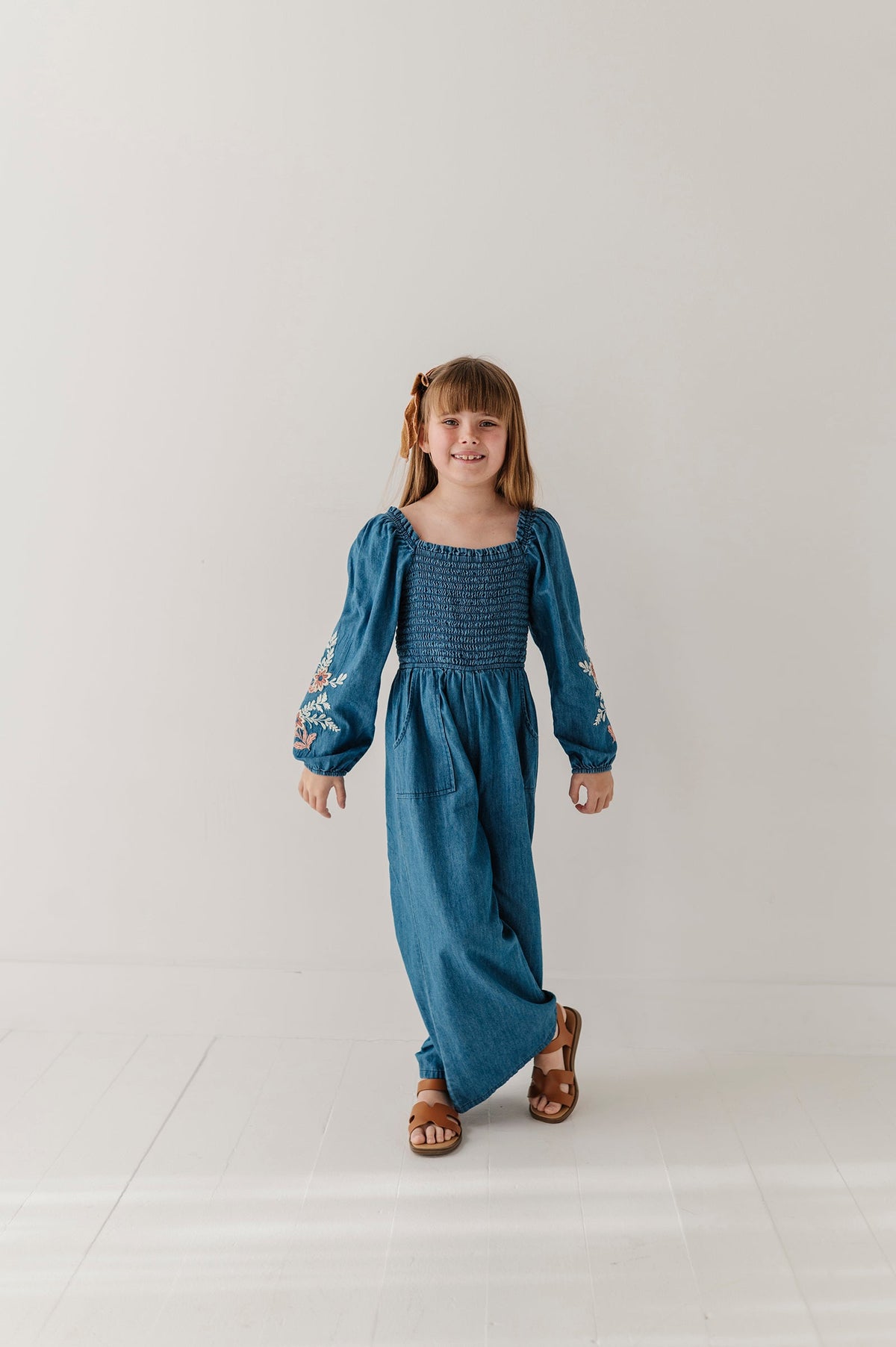 A young girl with light brown hair in pigtails stands in a studio wearing a smocked blue embroidered chambray romper with long puff sleeves and embroidered details on the cuffs, paired with brown sandals.