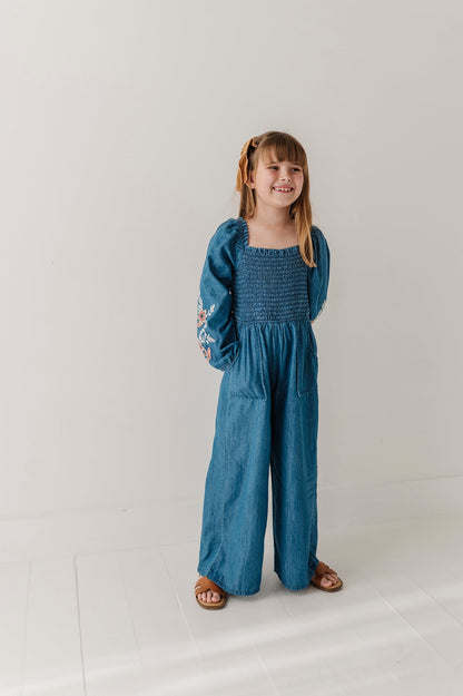 A young girl with blonde hair in pigtails stands in a studio wearing a blue chambray smocked jumpsuit with wide legs, puffed sleeves, and embroidered details on one sleeve, paired with brown sandals.