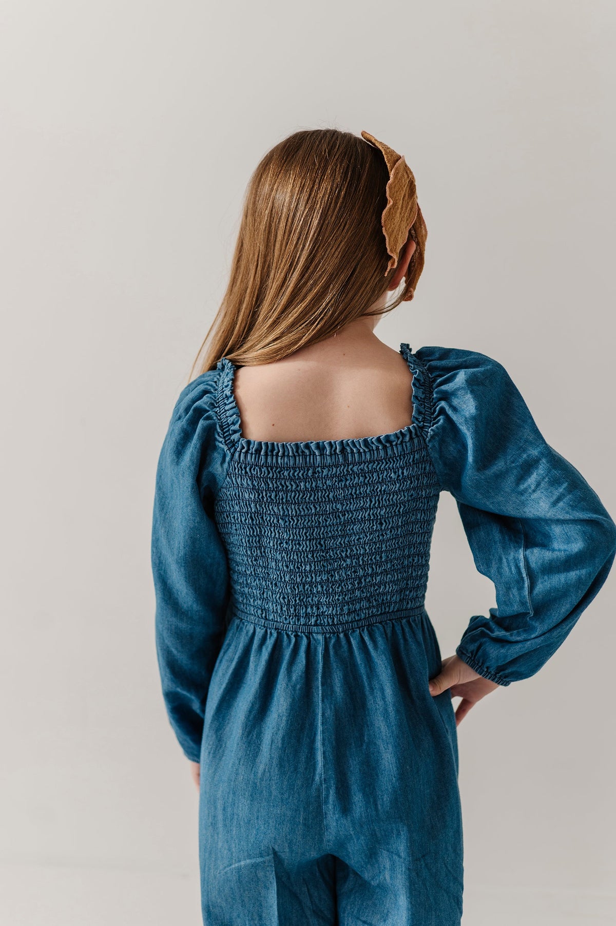 A child wearing a smocked blue denim-like chambray jumpsuit, shown from the back against a plain light gray studio background.