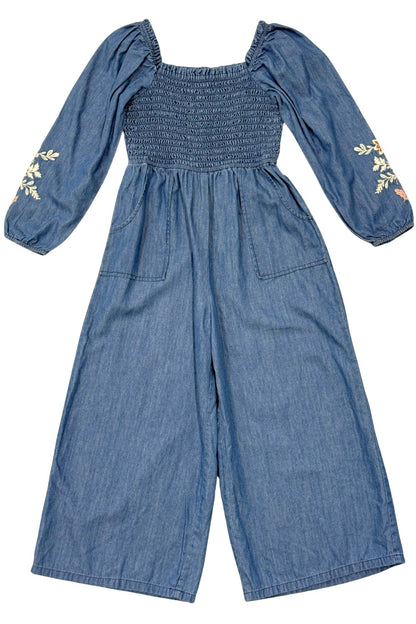 A blue chambray denim jumpsuit for girls, featuring a smocked bodice, square neckline, puff sleeves with floral embroidery on the cuffs, and wide legs, shown against a plain white background.