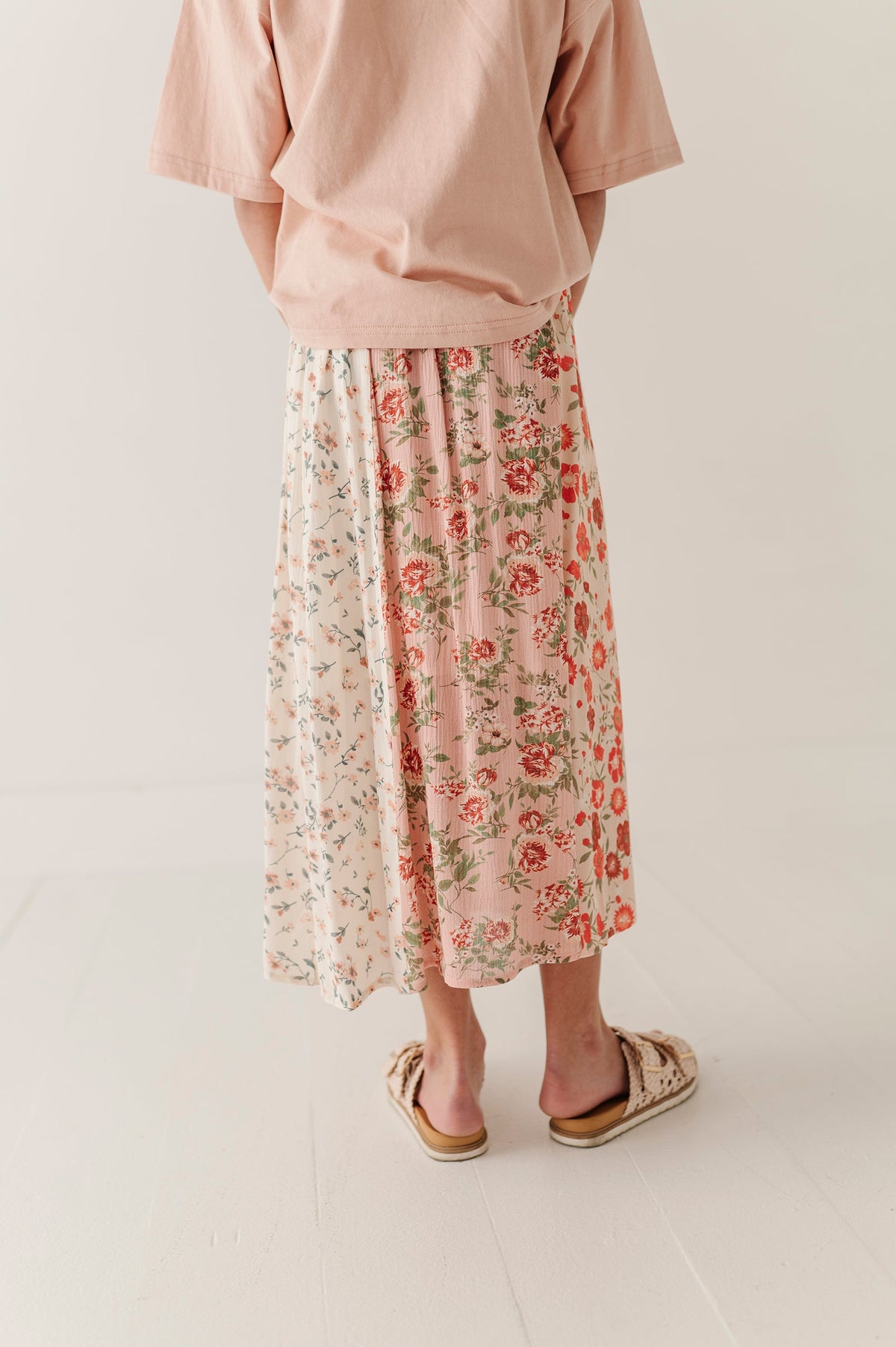 A person shown from the back wearing a dusty pink short-sleeve top and a midi skirt with a pink, red, and green floral garden pattern. They are wearing beige slide sandals on a clean, light-colored studio floor.
