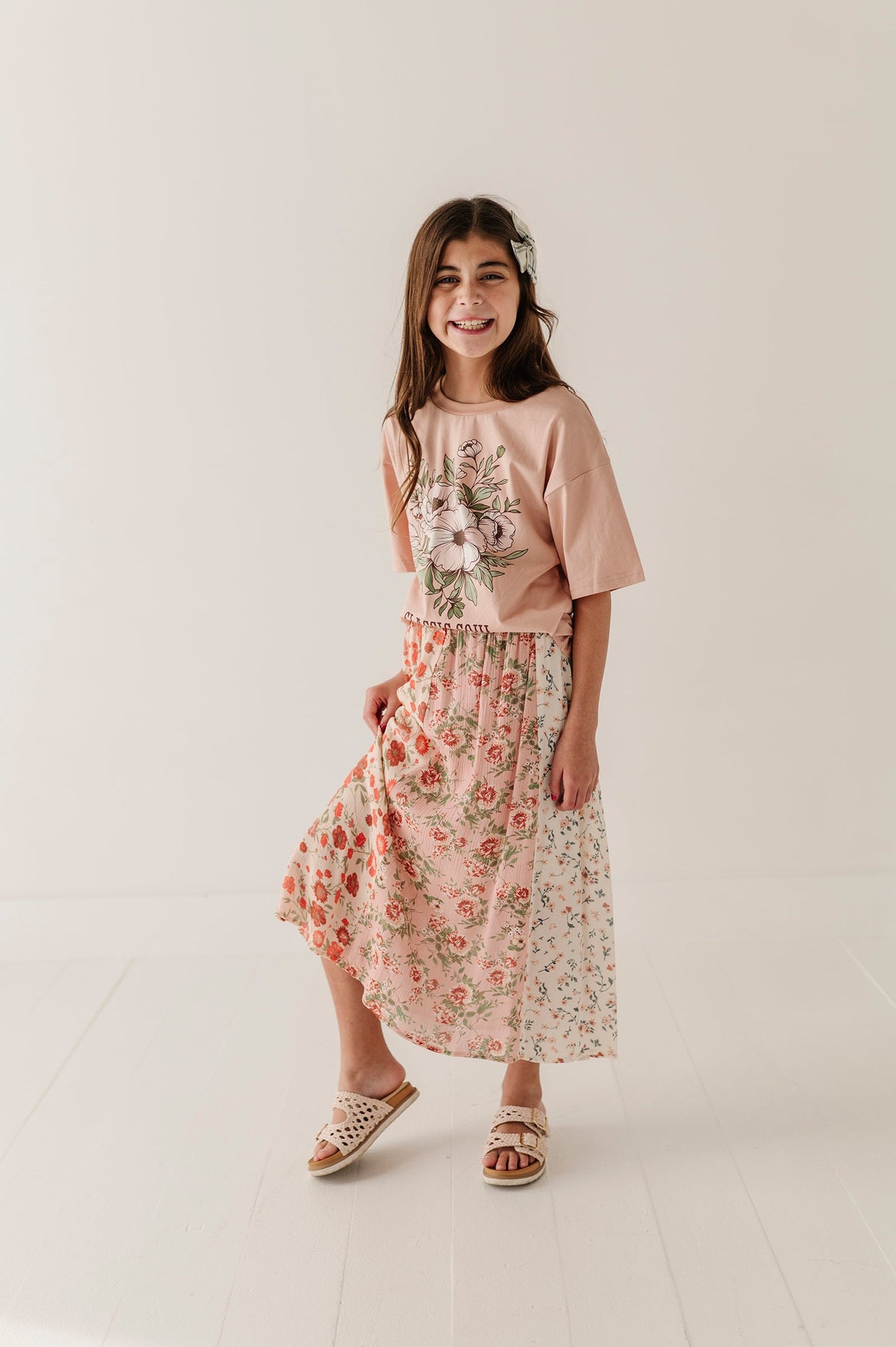 A young girl modeling a two-piece outfit consisting of a dusty pink short-sleeve top with a floral graphic and a matching multirose midi skirt, paired with woven sandals, in a bright studio setting.