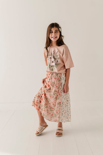 A young girl modeling a two-piece outfit consisting of a dusty pink short-sleeve top with a floral graphic and a matching multirose midi skirt, paired with woven sandals, in a bright studio setting.