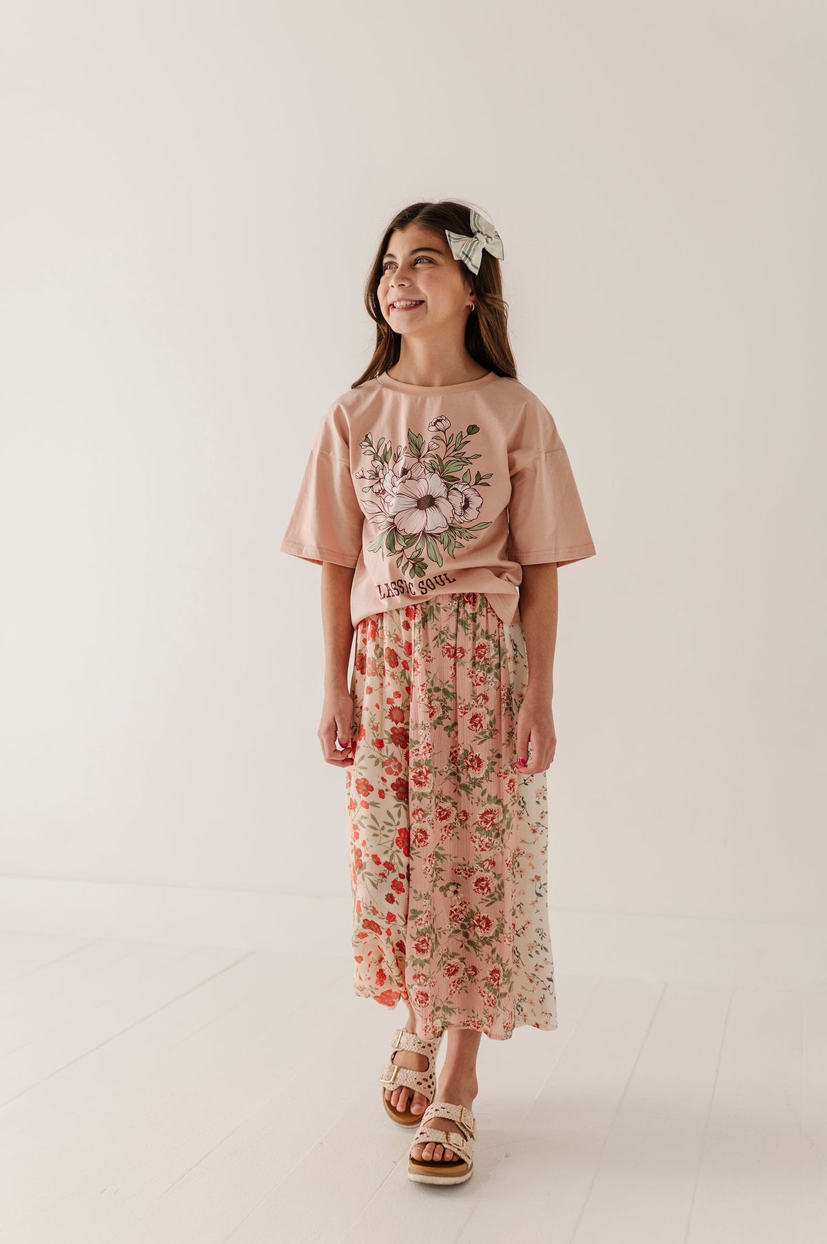 A young girl wearing a dusty pink short-sleeve t-shirt with a floral graphic and a matching floral multirose midi skirt, standing against a plain off-white studio background.