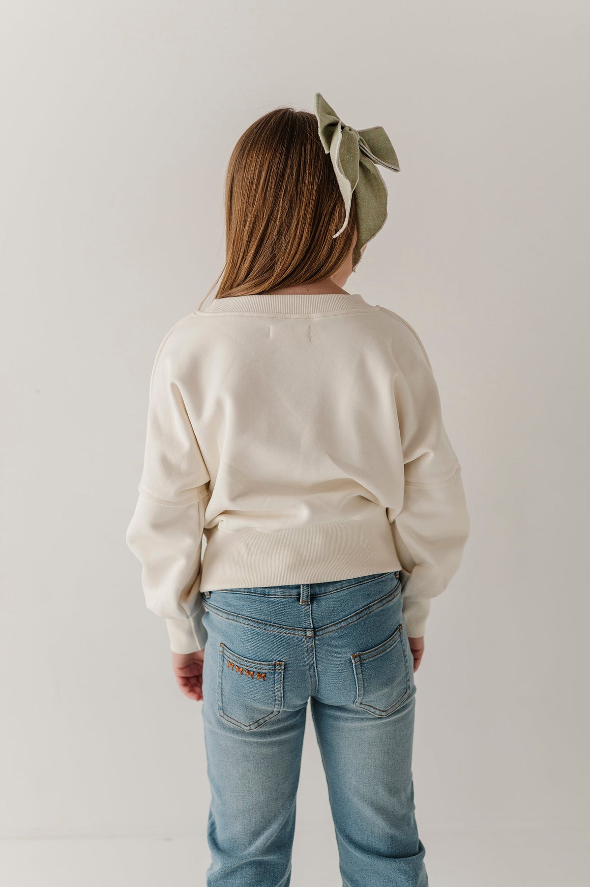 A child viewed from behind wearing a cream-colored long-sleeve sweatshirt and light blue jeans, with a sage green bow headband. The photo is taken against a plain, light off-white studio background.