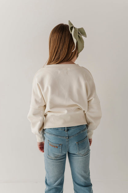 A child viewed from behind wearing a cream-colored long-sleeve sweatshirt and light blue jeans, with a sage green bow headband. The photo is taken against a plain, light off-white studio background.
