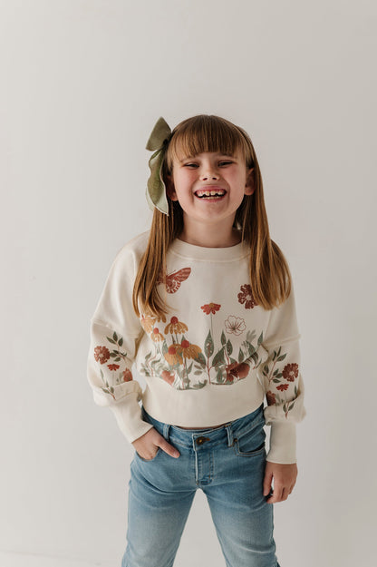 Young girl wearing a drop shoulder floral garden sweatshirt and blue jeans with a white background