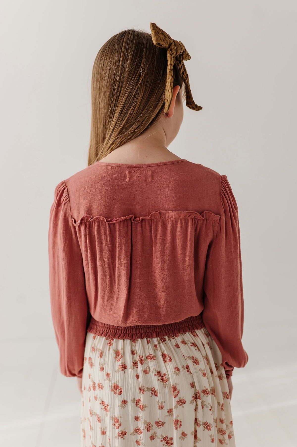 A child viewed from behind wearing a dusty rose long-sleeve blouse with ruffled detailing across the upper back and a cream floral skirt against a plain light background.