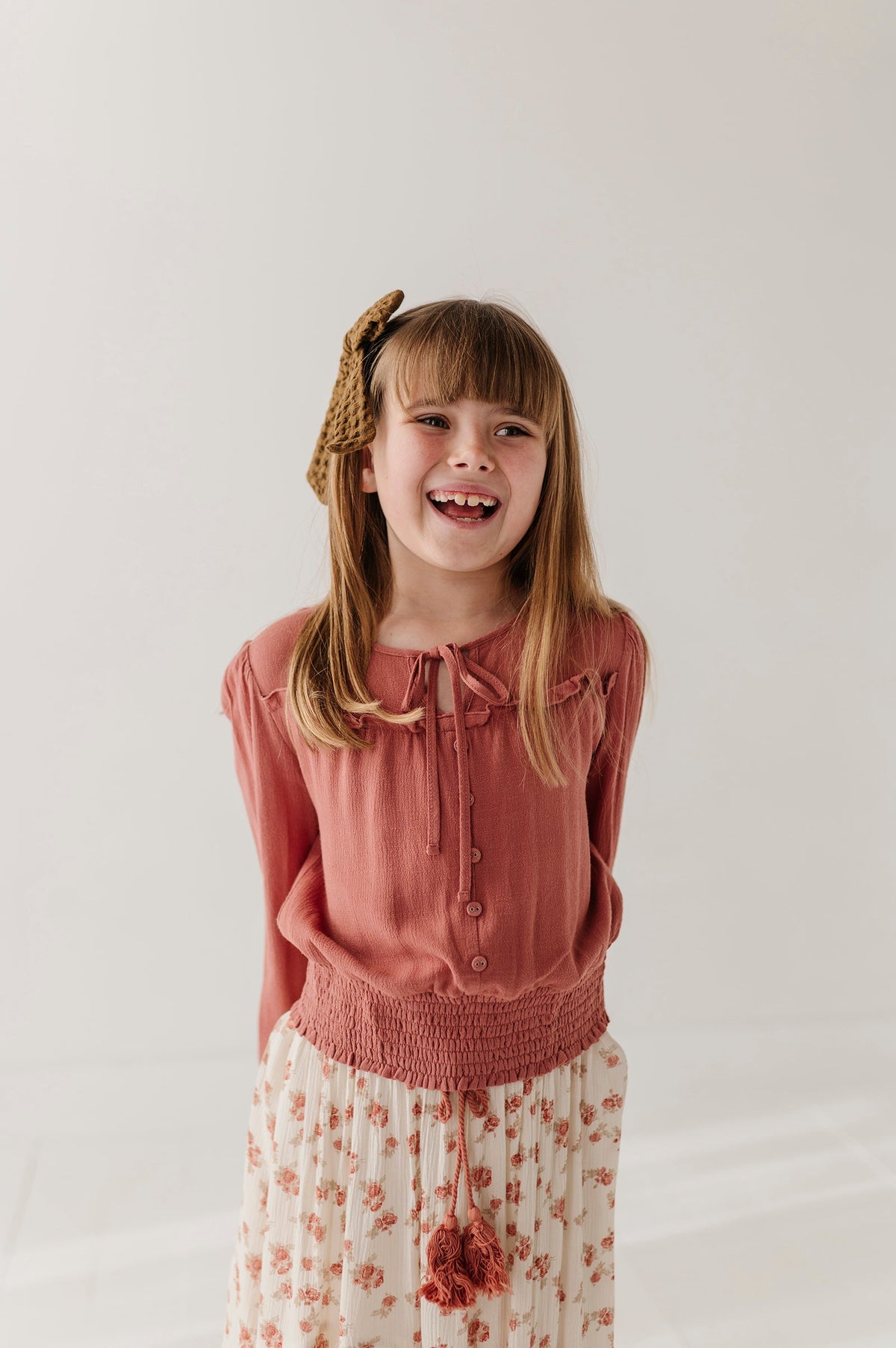 A young girl with blonde hair wearing a brick dust colored  long-sleeve button-up top and a cream floral skirt, smiling in a studio setting.