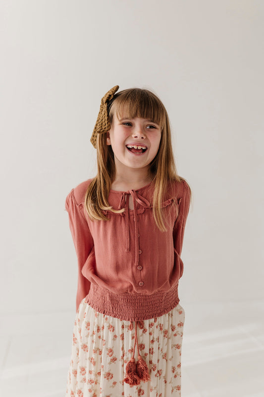 A young girl with blonde hair wearing a brick dust colored  long-sleeve button-up top and a cream floral skirt, smiling in a studio setting.