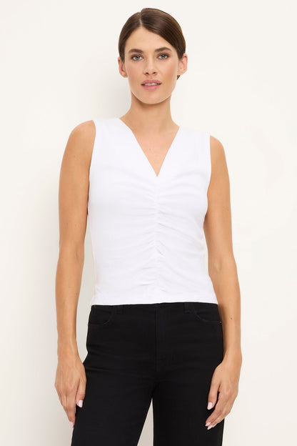 A woman modeling a white sleeveless V-neck Lune top with ruched detailing on the front, paired with black pants, against a plain off-white studio background.