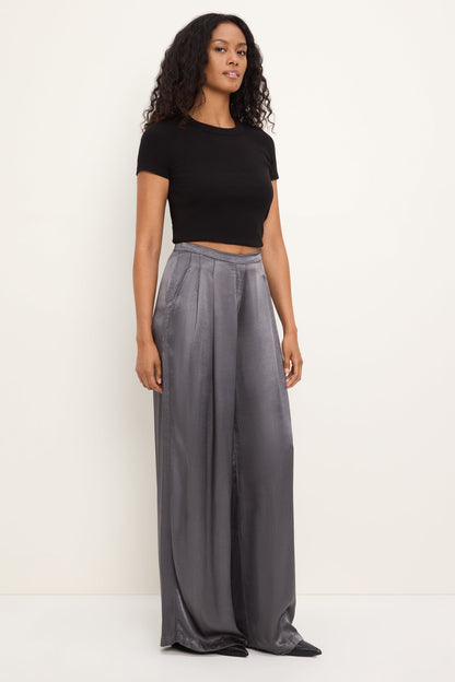 A woman modeling a black short-sleeve crop top and high-waisted, wide-leg thunder gray satin pants against a plain off-white studio background.