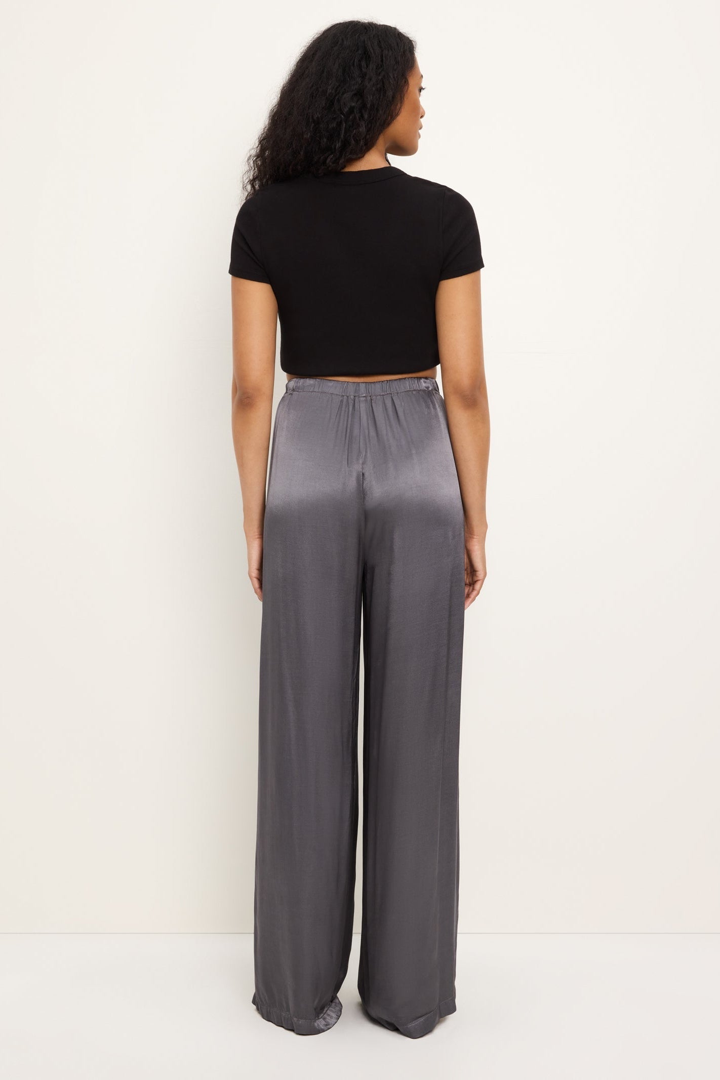 A woman shown from the back wearing a black short-sleeve crop top and high-waisted, wide-leg thunder gray satin pants against a plain off-white studio background.