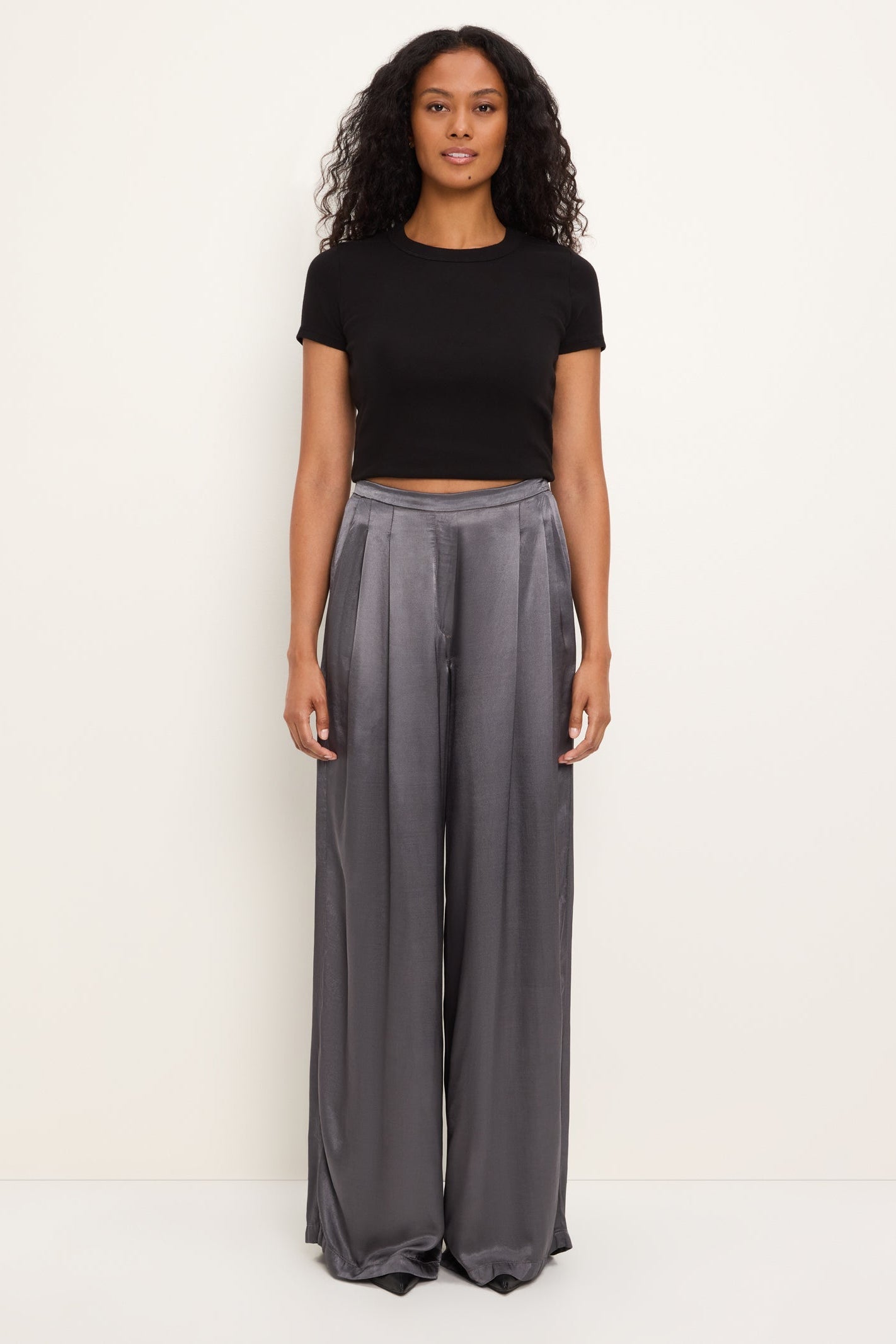 A woman stands against a plain off-white studio background wearing a black short-sleeve cropped T-shirt and high-waisted, wide-leg thunder gray satin pants.