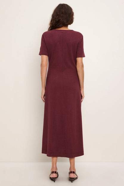 A woman viewed from the back wearing a short-sleeve, knee-length port royale colored column dress and black pointed-toe flats against a plain off-white studio background.