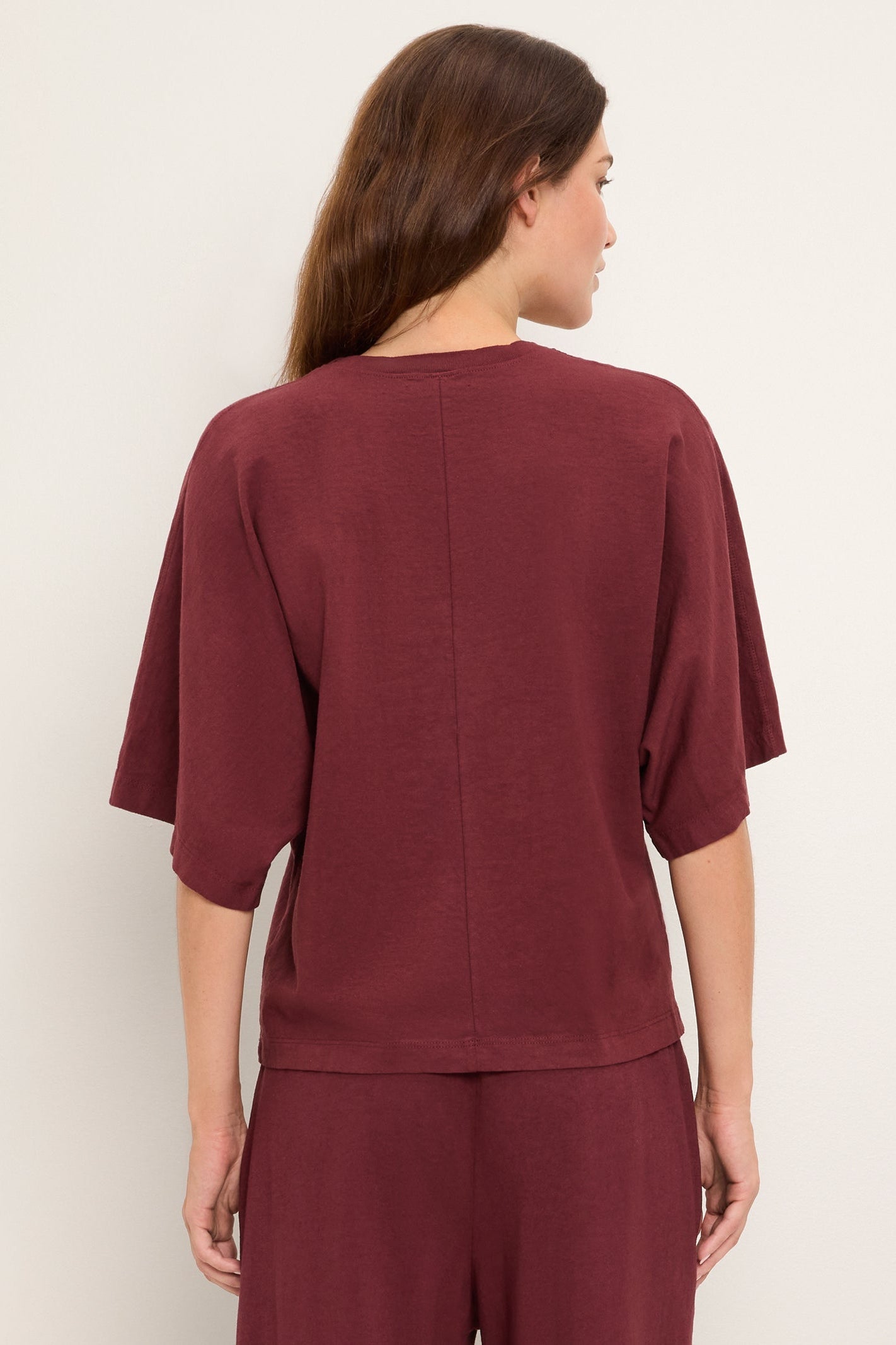 A woman shown from the back wearing a loose-fitting short-sleeve top and matching pants in a deep burgundy port royale color, photographed against a plain light neutral background.
