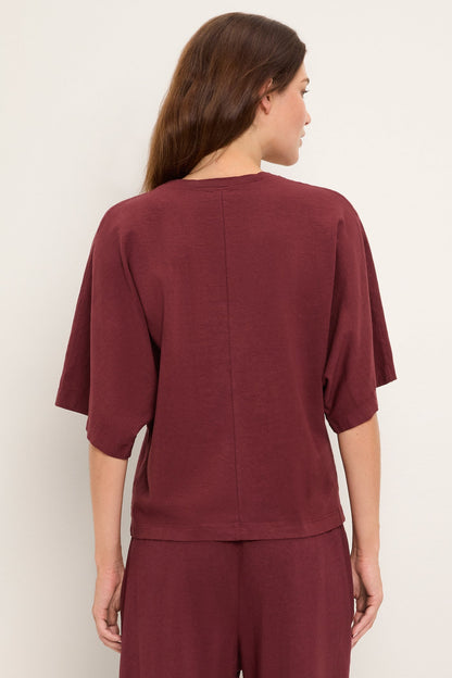 A woman shown from the back wearing a loose-fitting short-sleeve top and matching pants in a deep burgundy port royale color, photographed against a plain light neutral background.