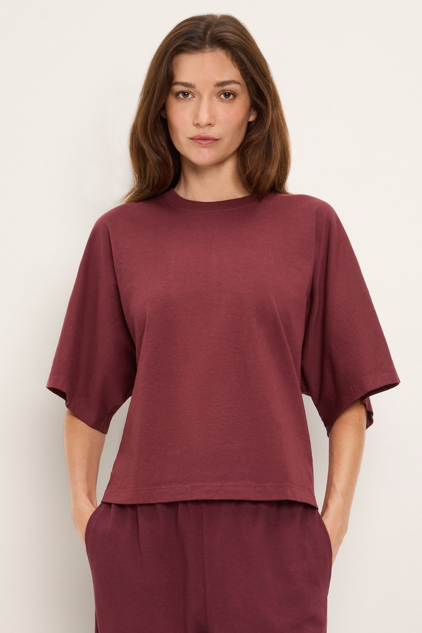A woman modeling a loose-fitting short-sleeve crewneck top in a port royale burgundy shade, paired with matching pants, against a plain off-white studio background.