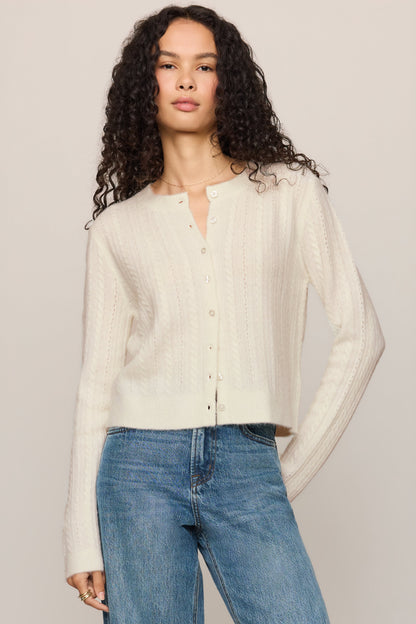 A woman with long curly dark hair wearing a 'cloud' cream-colored, textured long-sleeve button-up cardigan and blue jeans, standing against a solid light beige background.