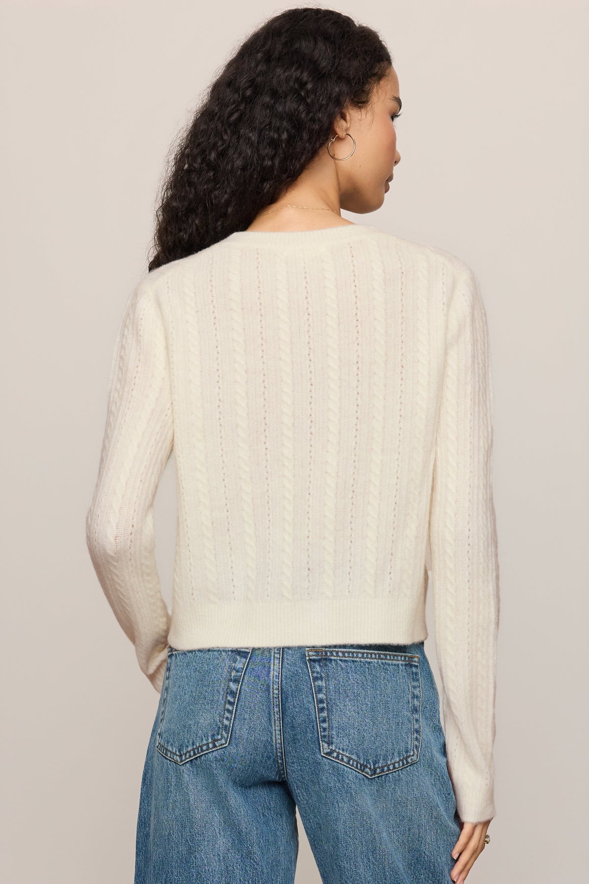 A woman shown from the back wearing a 'cloud' cream-colored ribbed knit sweater and blue jeans against a plain light beige studio background.