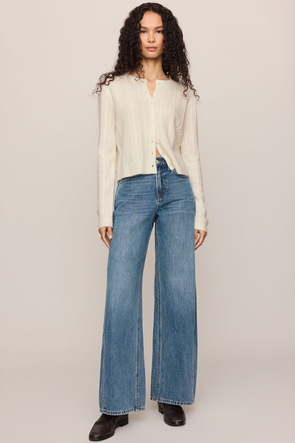 A model stands against a plain light beige studio background wearing a 'cloud' cream ribbed knit long-sleeve cardigan, blue wide-leg jeans, and black shoes.