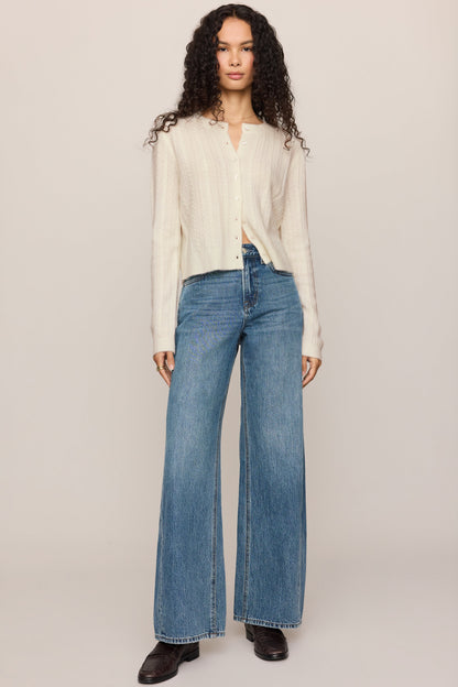 A model stands against a plain light beige studio background wearing a 'cloud' cream ribbed knit long-sleeve cardigan, blue wide-leg jeans, and black shoes.