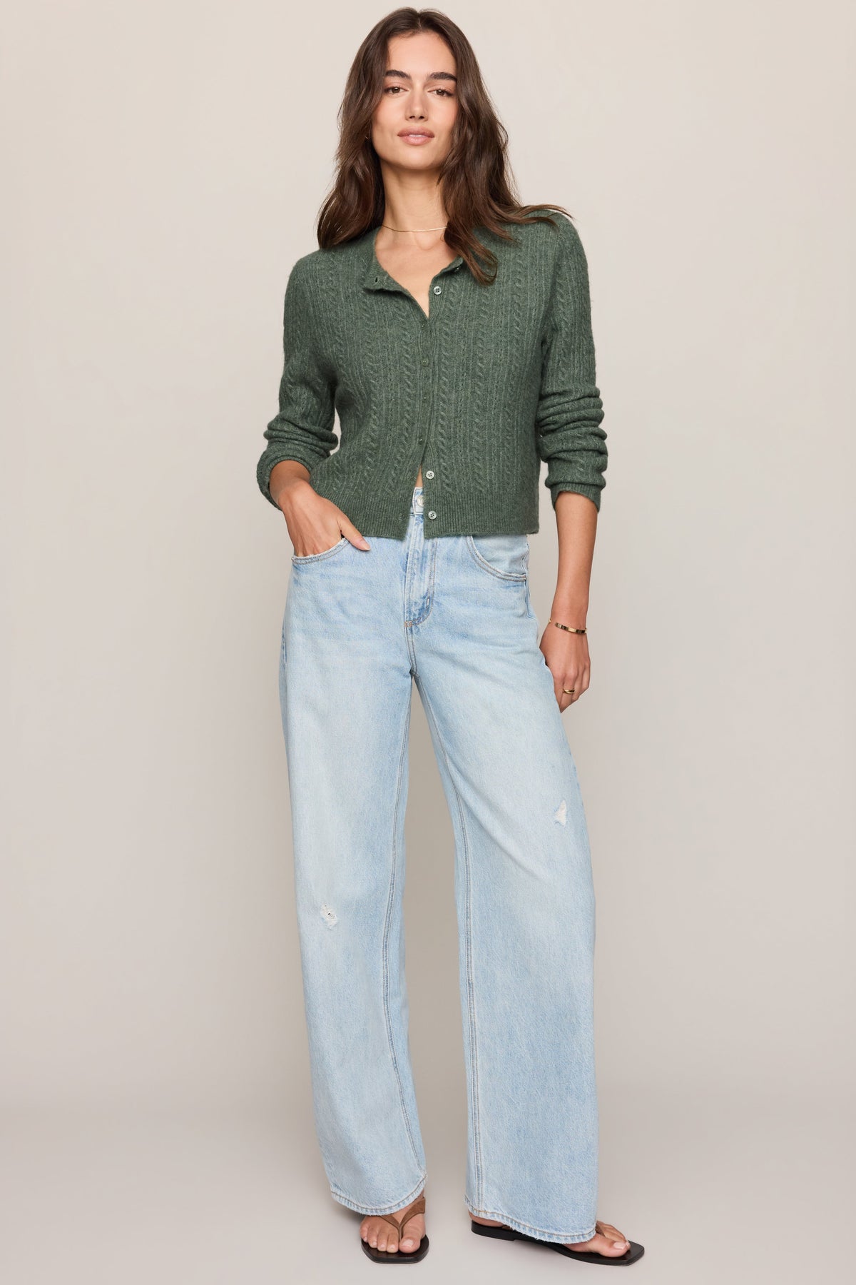 A woman modeling a monstera dark green textured knit cardigan and light-wash wide-leg jeans against a solid beige studio background.