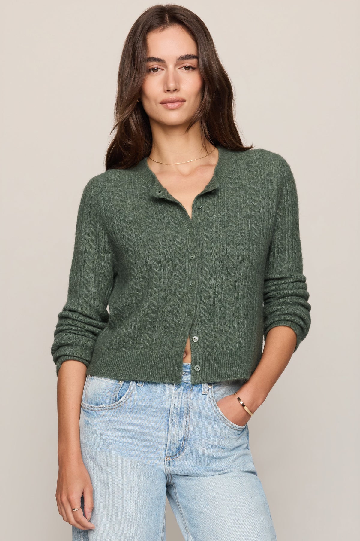A woman modeling a monstera dark green cable-knit button-up cardigan sweater paired with light-wash blue jeans, set against a plain beige studio background.