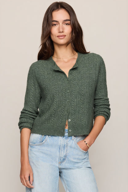 A woman modeling a monstera dark green cable-knit button-up cardigan sweater paired with light-wash blue jeans, set against a plain beige studio background.