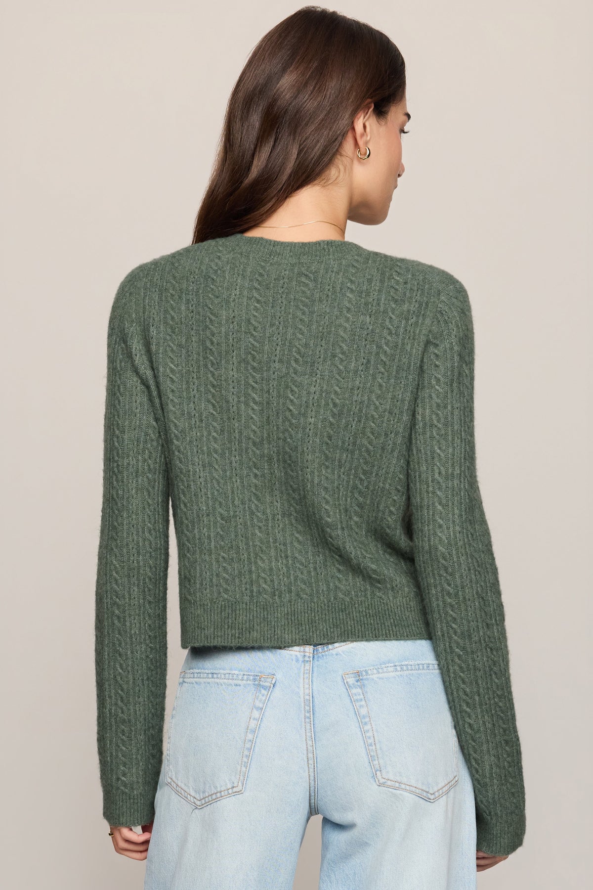 A woman viewed from the back wearing a sage green cable-knit cropped sweater and light-wash blue jeans against a plain beige studio background.