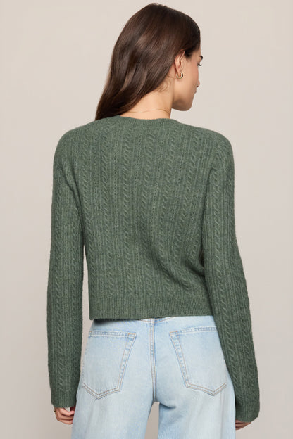 A woman viewed from the back wearing a sage green cable-knit cropped sweater and light-wash blue jeans against a plain beige studio background.