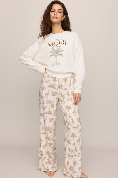 A woman modeling a matching two-piece lounge set featuring a cream sweatshirt with a 'SAFARI' palm tree graphic and wide-leg pants with a beige safari/palm tree print on a cream background.