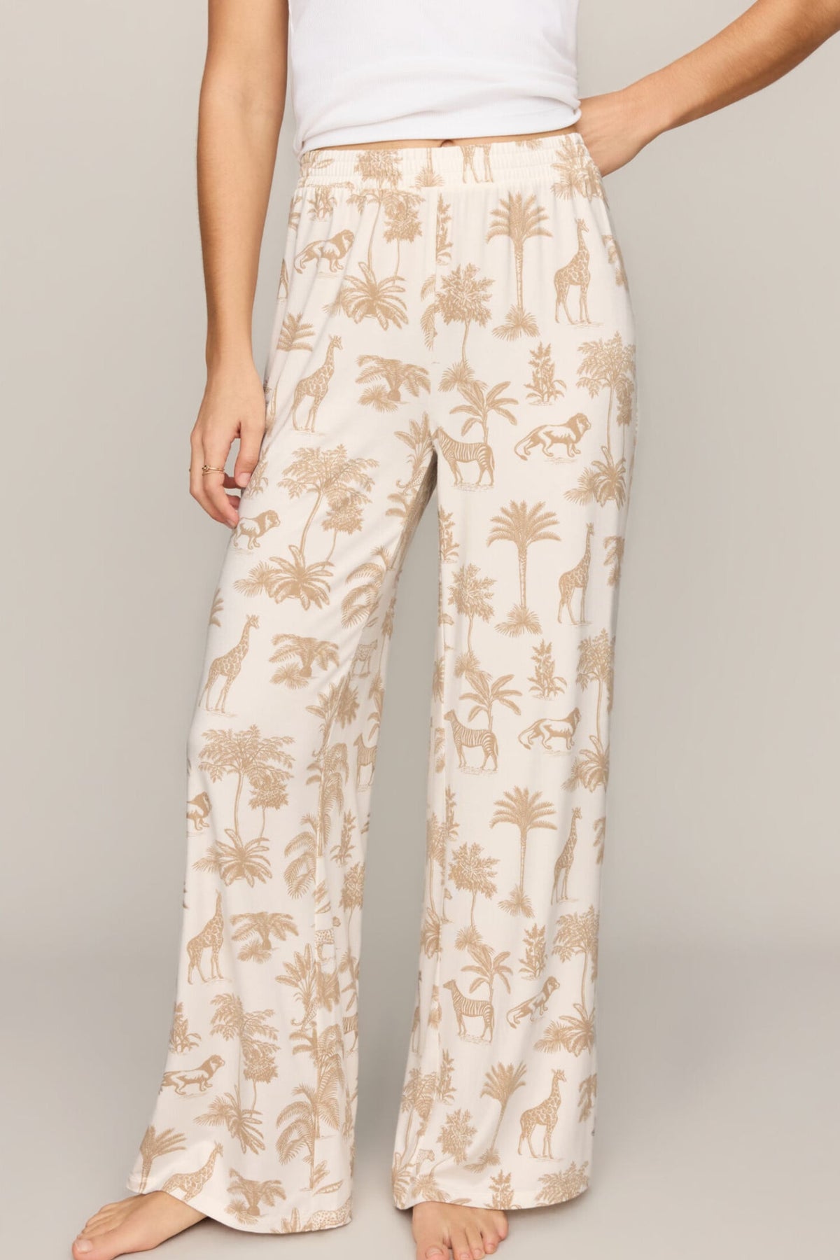 A person wearing loose, wide-leg pajama pants with an allover safari-style print of palm trees, giraffes, and zebras on a warm off-white background, paired with a white top against a plain beige studio backdrop.