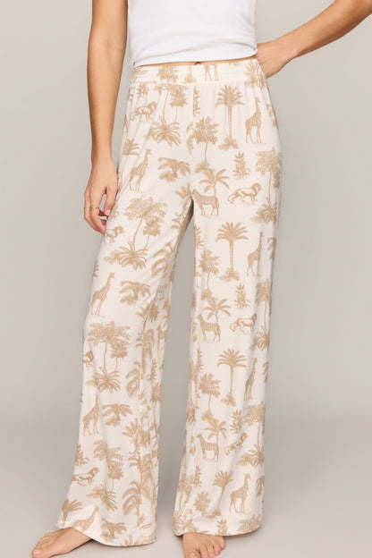 A person wearing loose, wide-leg pajama pants with an allover safari-style print of palm trees, giraffes, and zebras on a warm off-white background, paired with a white top against a plain beige studio backdrop.