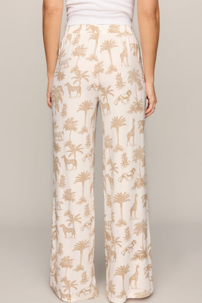 A person shown from the back wearing wide-leg pants with an allover safari-themed print of palm trees, giraffes, and zebras on a light cream background, paired with a white top against a neutral studio backdrop.