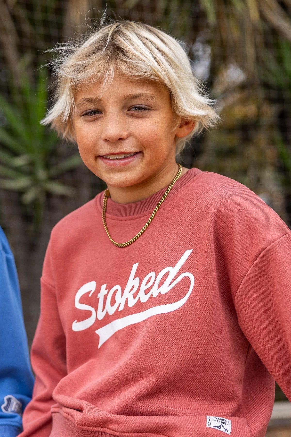 A young blonde boy wearing a dusty canyon red crewneck sweatshirt with 'Stoked' printed in white script across the chest, paired with a gold chain necklace. The photo is taken outdoors with a blurred natural background.