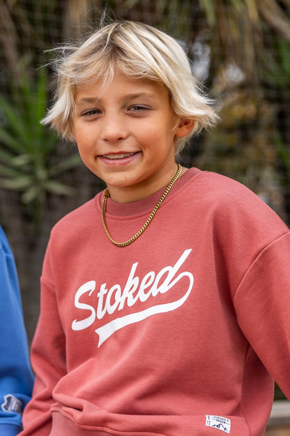 A young blonde boy wearing a dusty canyon red crewneck sweatshirt with 'Stoked' printed in white script across the chest, paired with a gold chain necklace. The photo is taken outdoors with a blurred natural background.