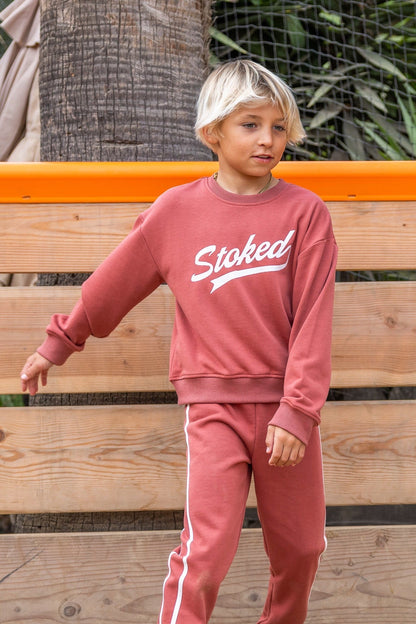 A young boy with blonde hair wearing a dusty rose sweatshirt and matching sweatpants with white side stripes and 'Stoked' written on the chest, standing in front of a wooden bench.