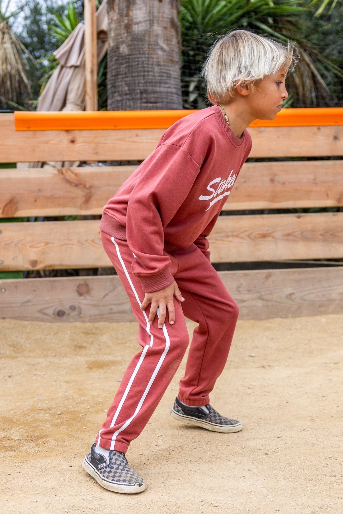 A young boy wearing a matching  canyon red sweatshirt and sweatpants set with white stripes, standing on a sandy ground in front of a wooden fence.