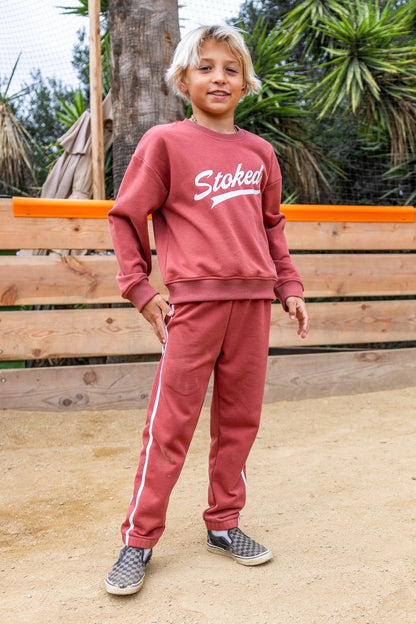 A child wearing a matching canyon red sweatshirt and sweatpants set with white script lettering on the chest and down the pants, standing outdoors in front of a wooden fence.