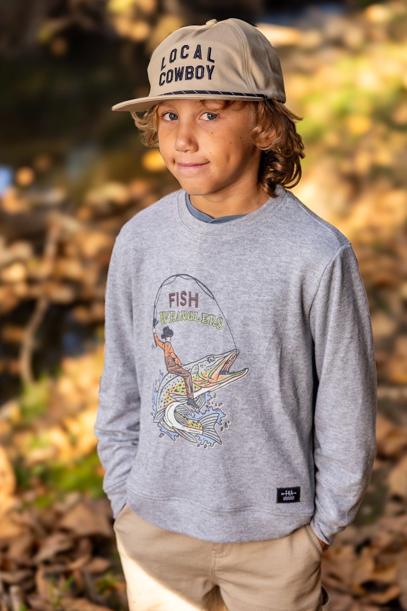 A young boy wearing a heather gray crewneck sweatshirt with a fish wrangler graphic on the front, khaki pants, and a beige 'LOCAL COWBOY' baseball cap, standing outdoors on a leaf-covered ground.